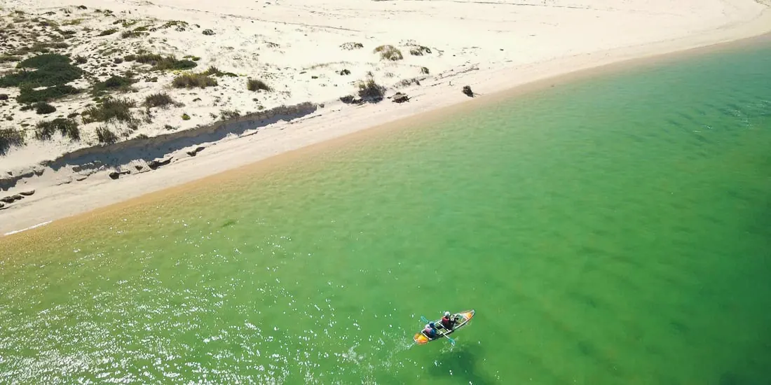 A clear kayak glides past a dune beach on the barrier island, green water fading to sand.