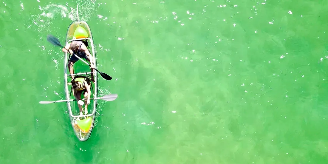 Seen from straight above, two paddlers in a transparent double kayak on bright green water.