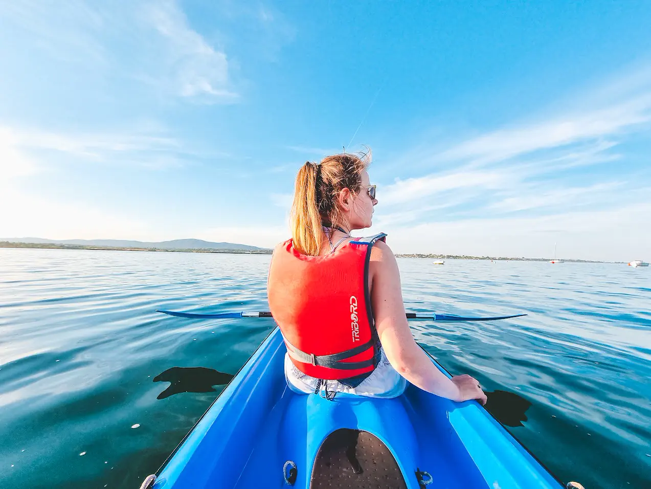 A paddler in a red life jacket sits in a blue kayak, looking across the still lagoon towards the hills.
