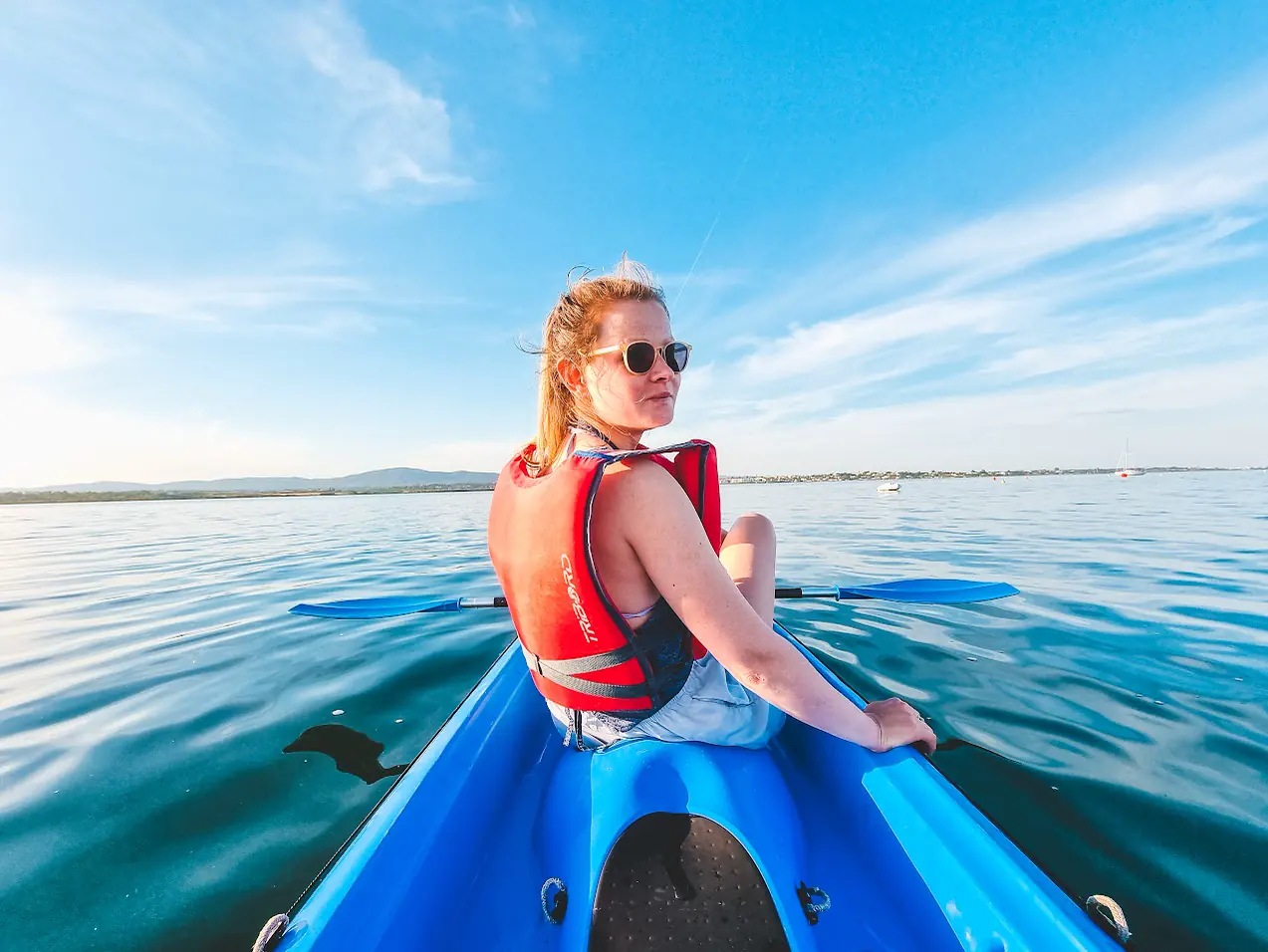 A smiling paddler in sunglasses and a red life jacket looks back from a blue kayak on the calm lagoon.